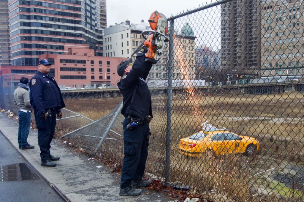 Police beside a yellow cab abandoned in a fenced lot